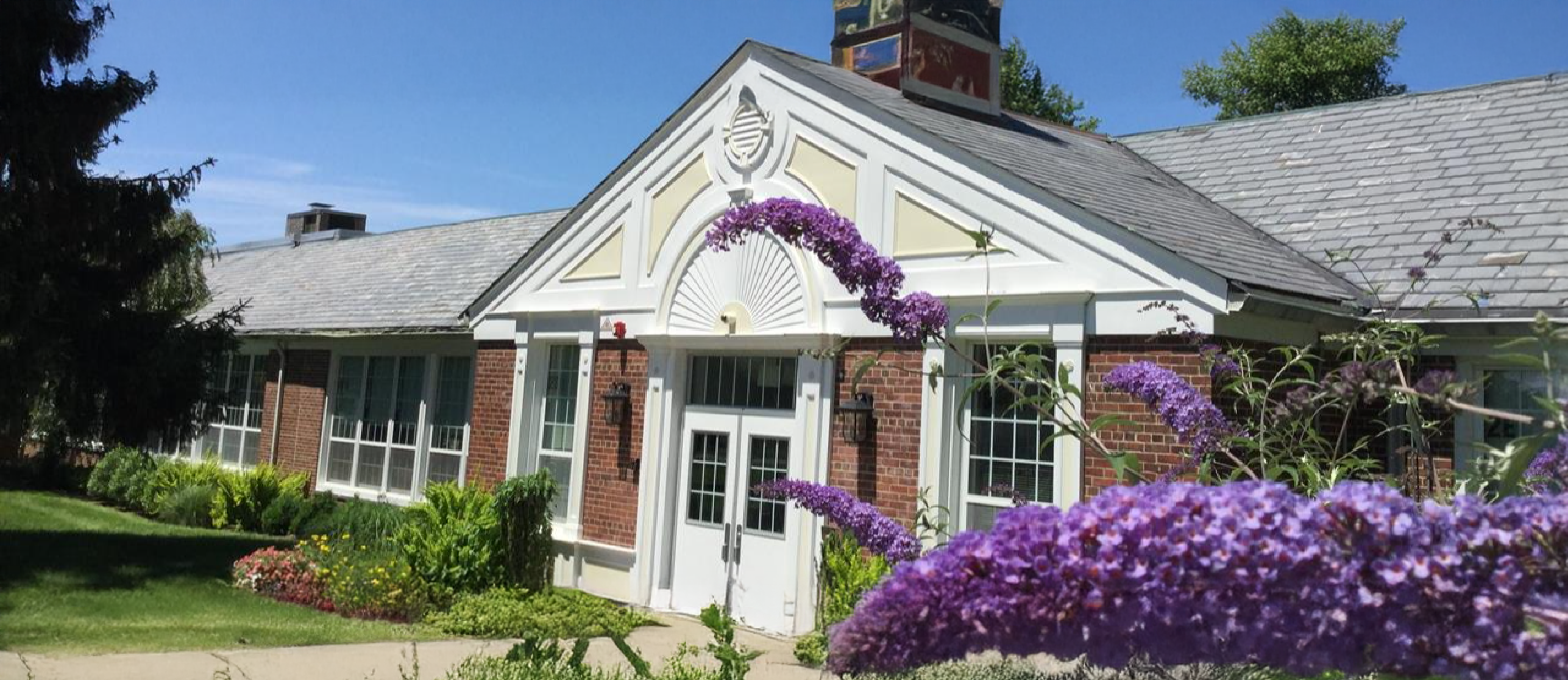 Exterior view of a brick school building with white trim and a central entrance, surrounded by greenery and purple flowering plants on a sunny day.
