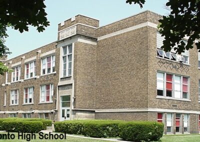 A brick building of a high school with large windows and a green entrance, surrounded by grass and trees.