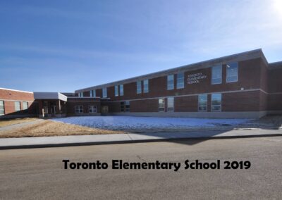 A view of Toronto Elementary School, showcasing its brick facade and large windows, with a clear blue sky above and snow on the ground in front.