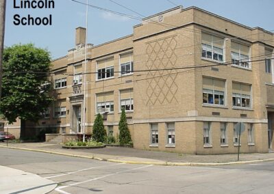 A brick building labeled 'Lincoln School' with multiple windows and greenery around it, set on a street corner.