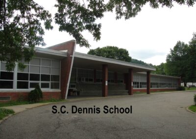 A view of the S.C. Dennis School building, featuring a long facade with large windows and a covered entrance, surrounded by trees.