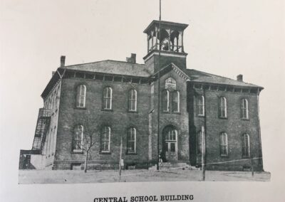 A historic brick school building with a clock tower and a flagpole, featuring multiple windows and a central entrance.