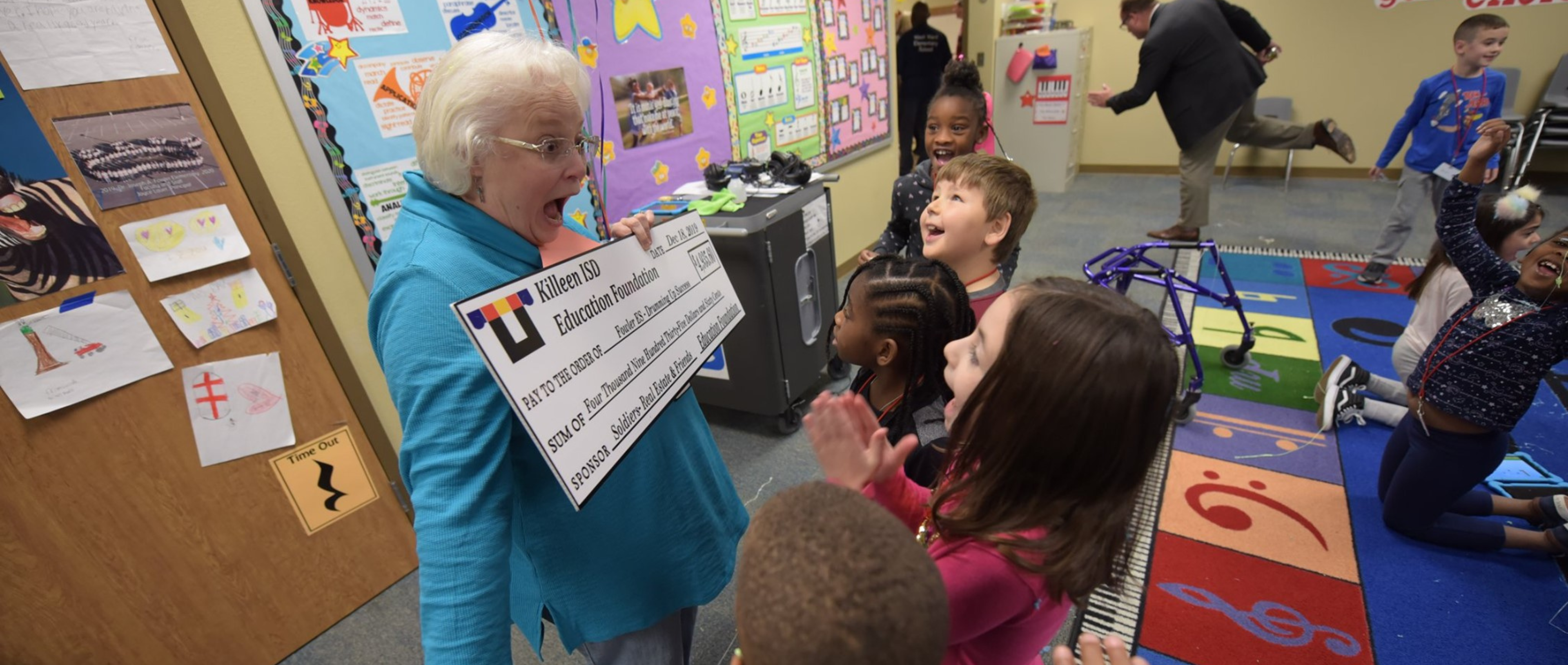 Teacher holding up a check with a surprised look on her face, children cheering as they see check