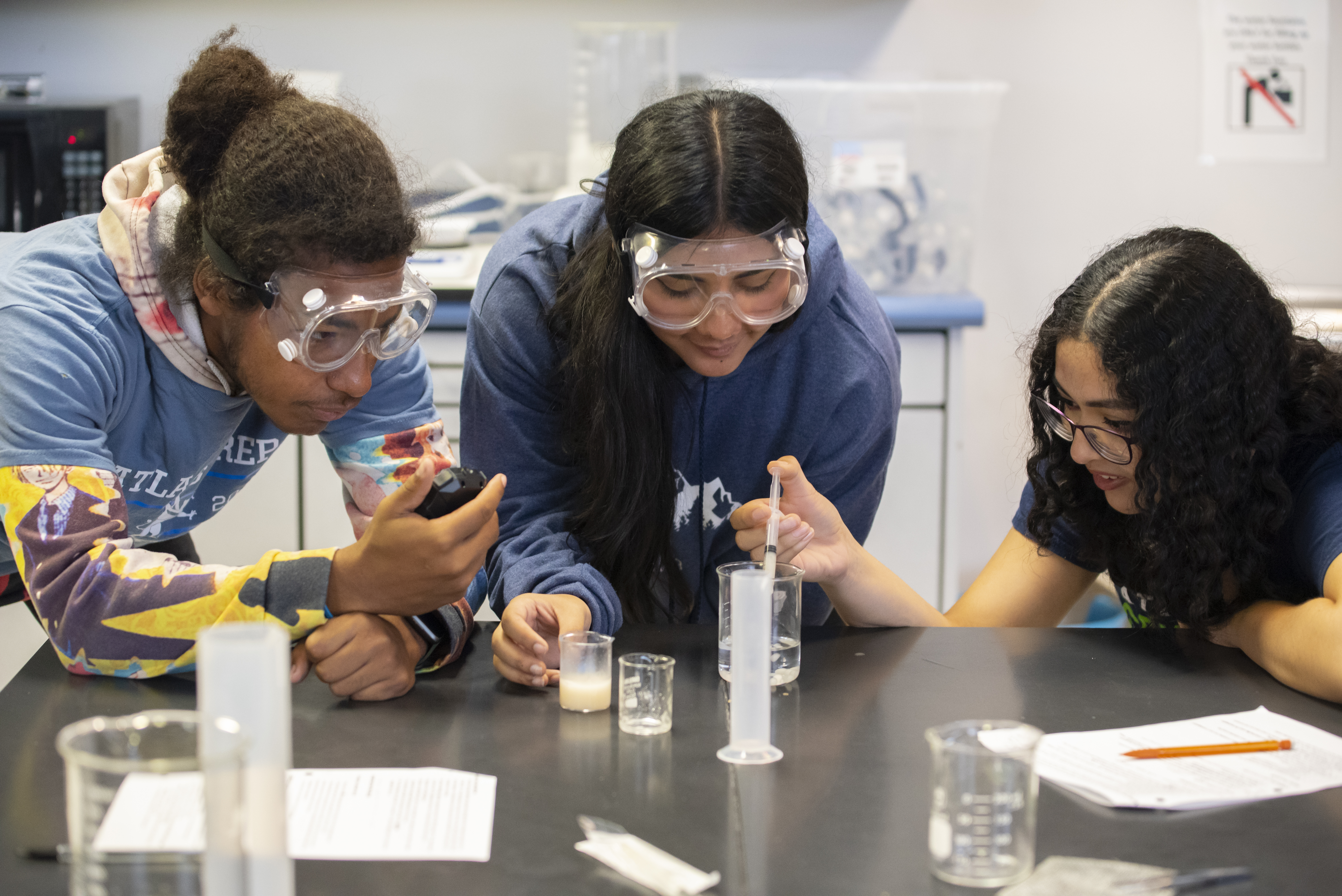 Three students wearing safety goggles are engaged in a science experiment, closely observing measurements in a graduated cylinder on a laboratory table.