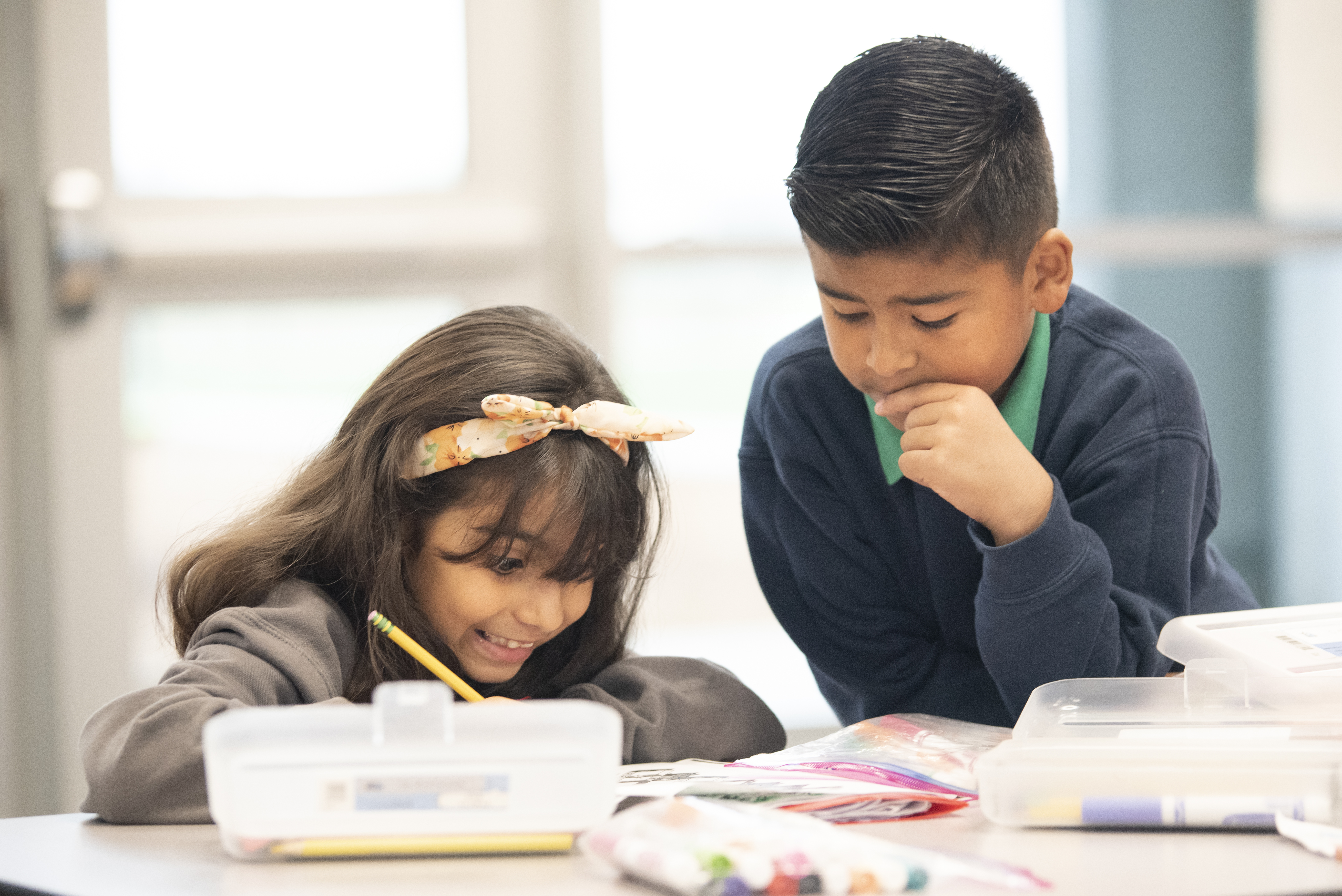 Two children, a girl with long hair and a headband and a boy with short hair, are engaged in a creative activity at a table, with various art supplies in front of them.