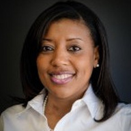 A woman with dark hair wearing a white shirt and a pearl necklace smiles at the camera against a dark background.