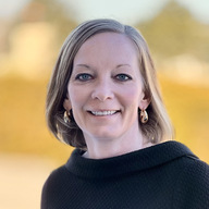 A woman with shoulder-length hair smiles warmly at the camera, wearing a black sweater and earrings, with a blurred outdoor background of soft colors.