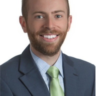 A smiling man with a beard, wearing a blue suit and a light green tie, poses for a professional headshot against a plain white background.