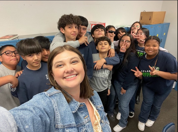 A group of students and a teacher pose together for a selfie, smiling and making playful gestures in a classroom setting.