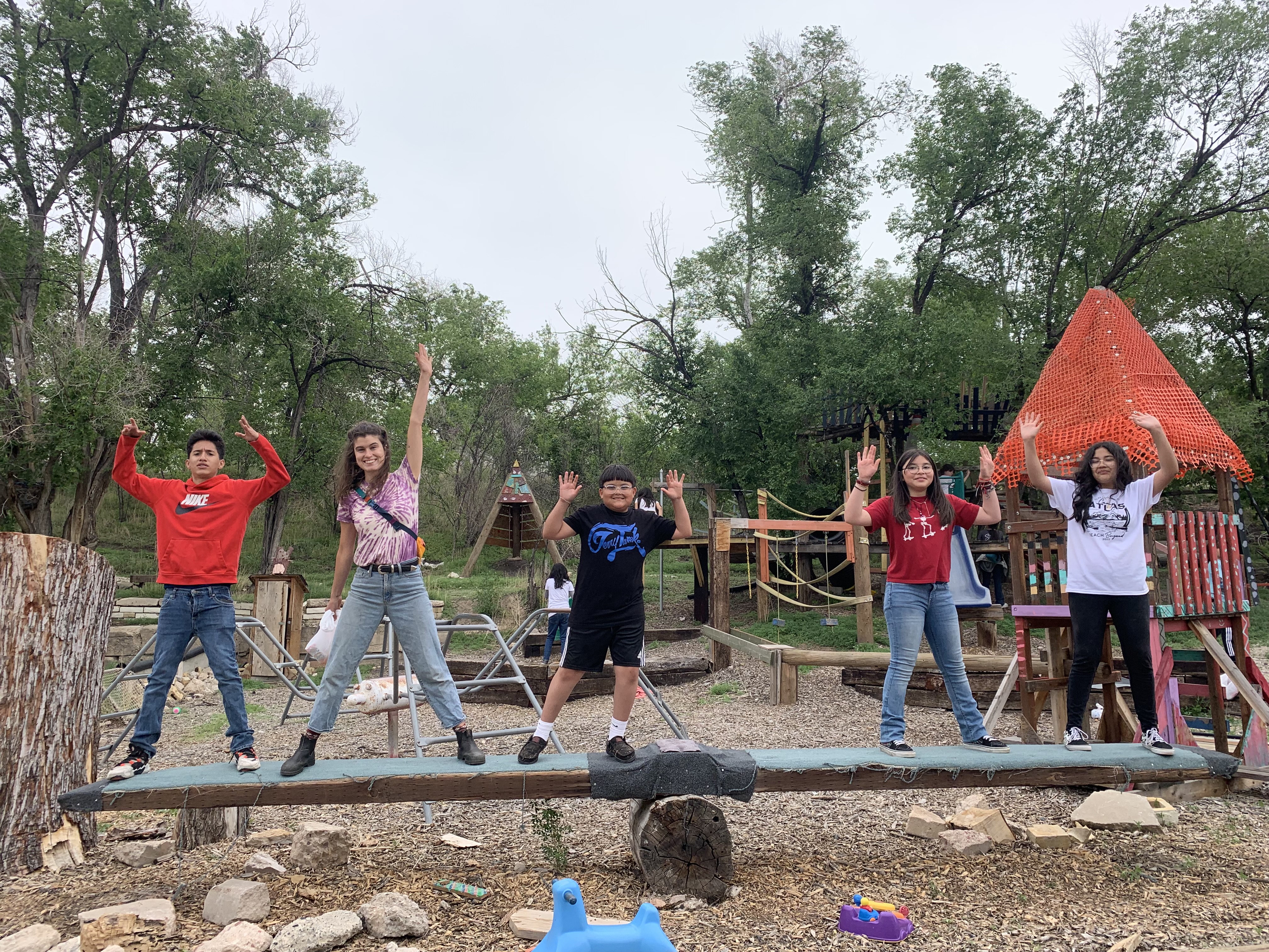 A group of five children stands on a balance beam in a playground, with their arms raised in celebration, surrounded by trees and play structures.