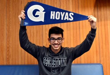 A smiling young man wearing glasses holds up a blue pennant that reads 'Hoyas' in white letters, while wearing a Georgetown University sweatshirt.