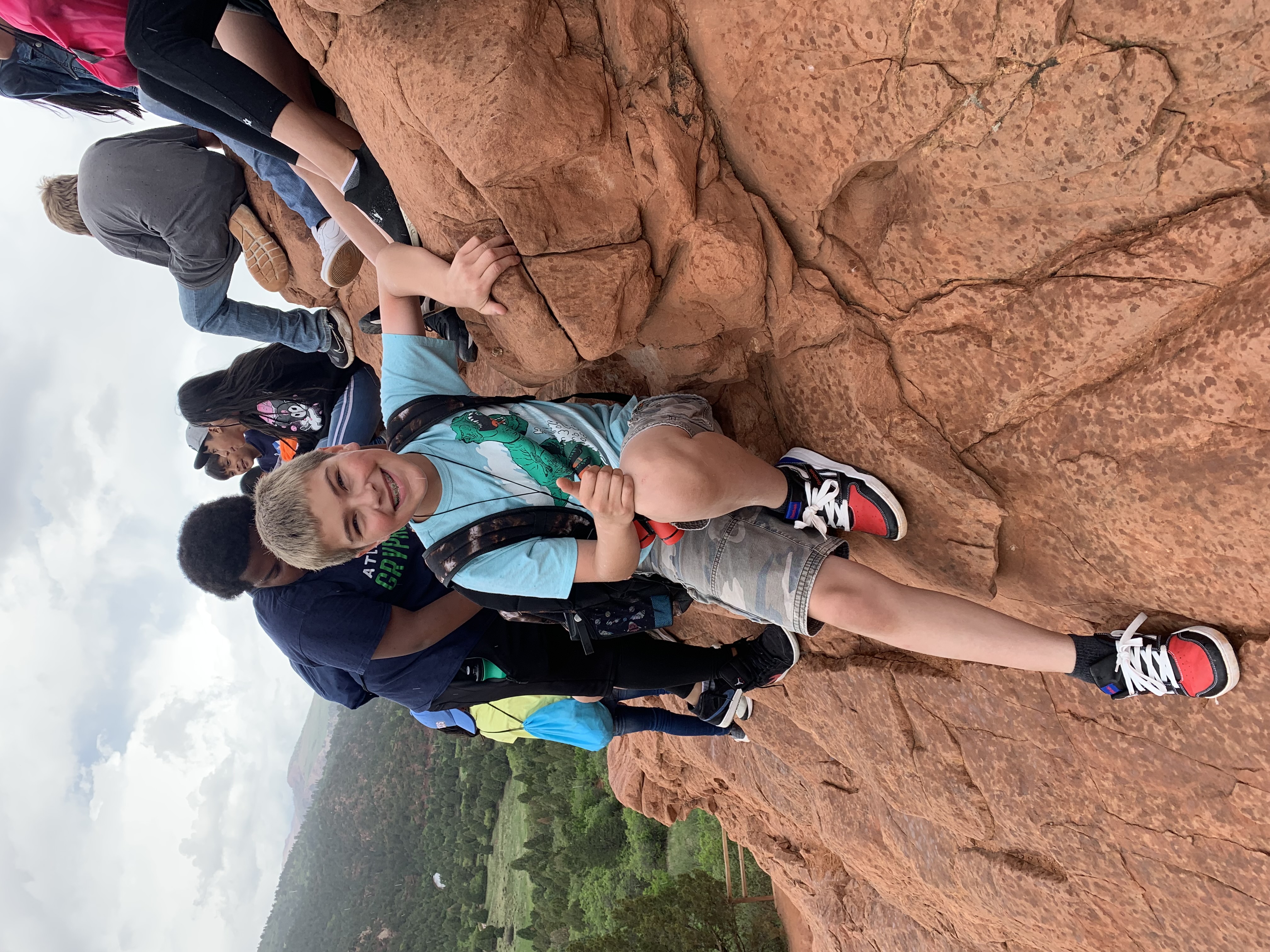 A young boy with a backpack sits on a rocky surface, smiling and giving a thumbs-up, while other children are climbing nearby in a scenic outdoor setting.