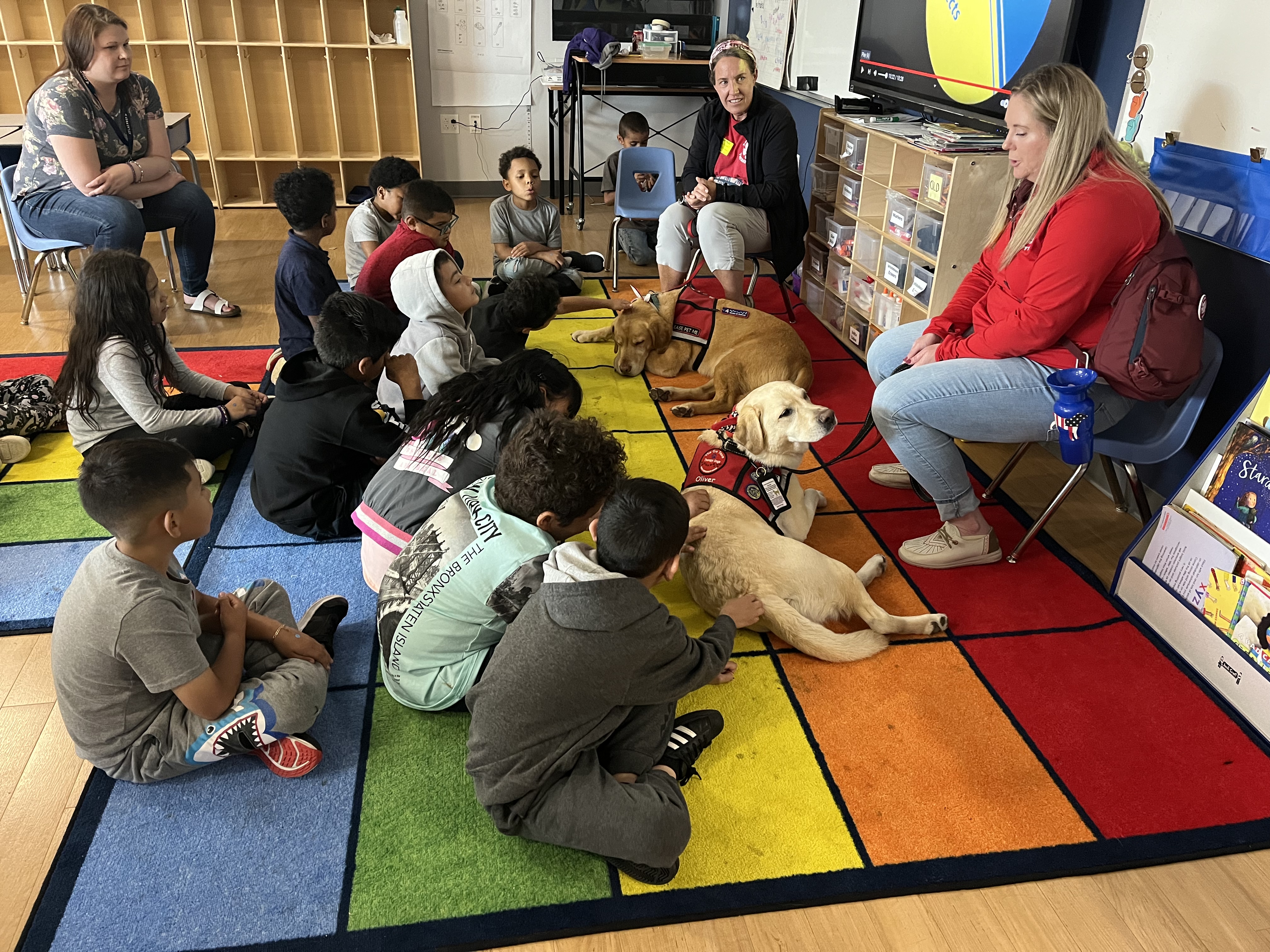 A group of children sitting on a colorful rug in a classroom, attentively listening to two adults, one of whom is holding a dog, while another dog lies on the floor nearby.