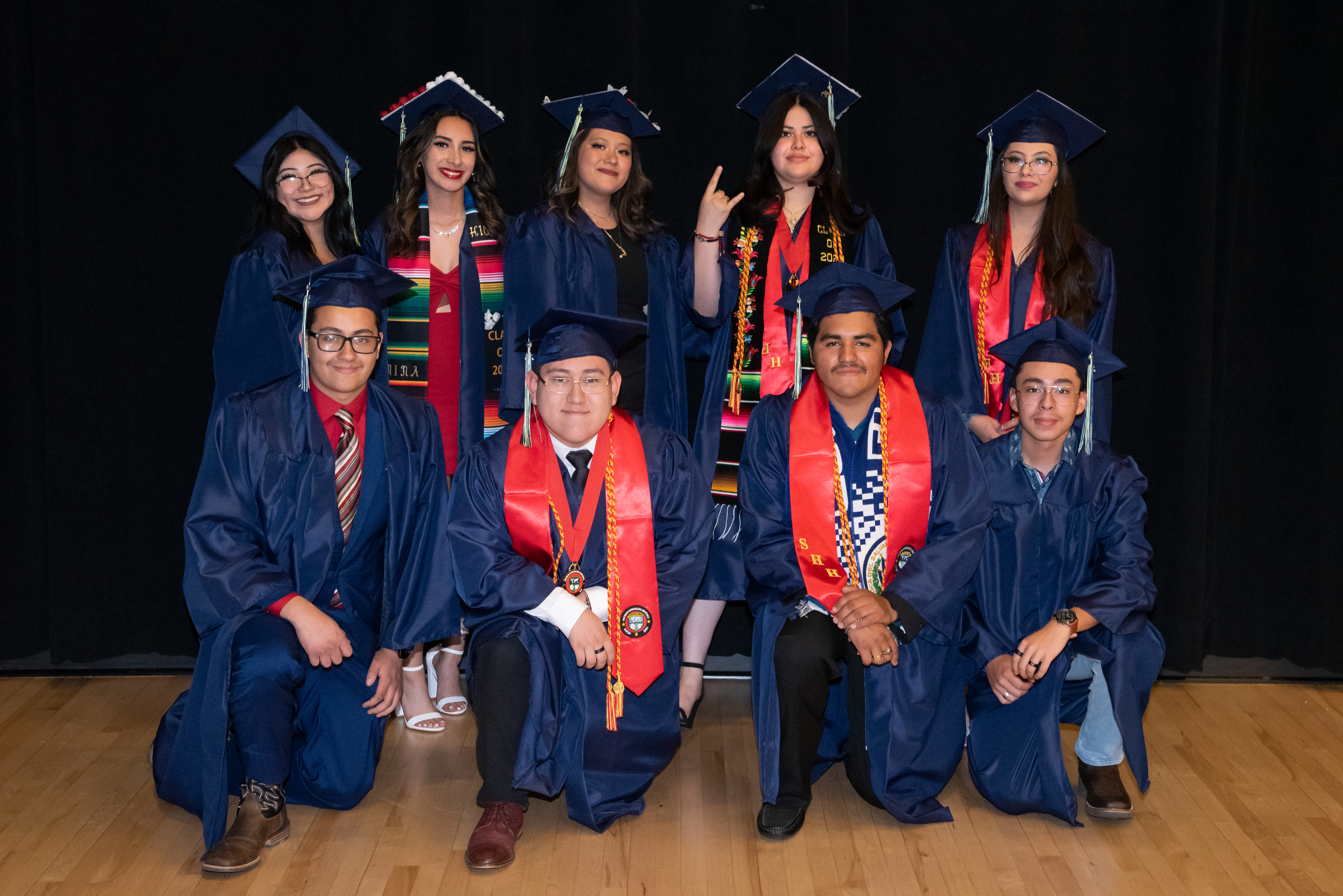 A group of nine graduates in blue caps and gowns, posing together with smiles and celebratory gestures, against a dark background.