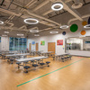 A spacious cafeteria featuring multiple tables and benches, with colorful circular decorations on the ceiling and large windows allowing natural light to enter.