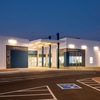 A modern building with a white exterior and blue accents, illuminated at dusk, featuring a covered entrance and parking area.