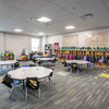 A bright and spacious classroom featuring several round tables, colorful storage cubbies, and a carpeted floor, designed for young learners.