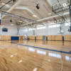 A spacious indoor basketball court featuring polished wooden floors, basketball hoops, and ample natural light from large windows.