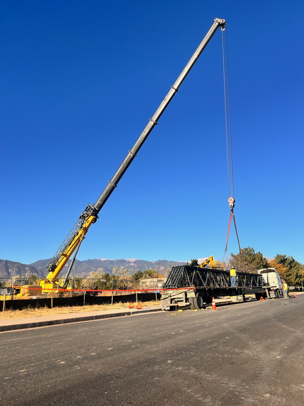 A large crane is lifting a heavy load on a construction site under a clear blue sky, with mountains visible in the background.