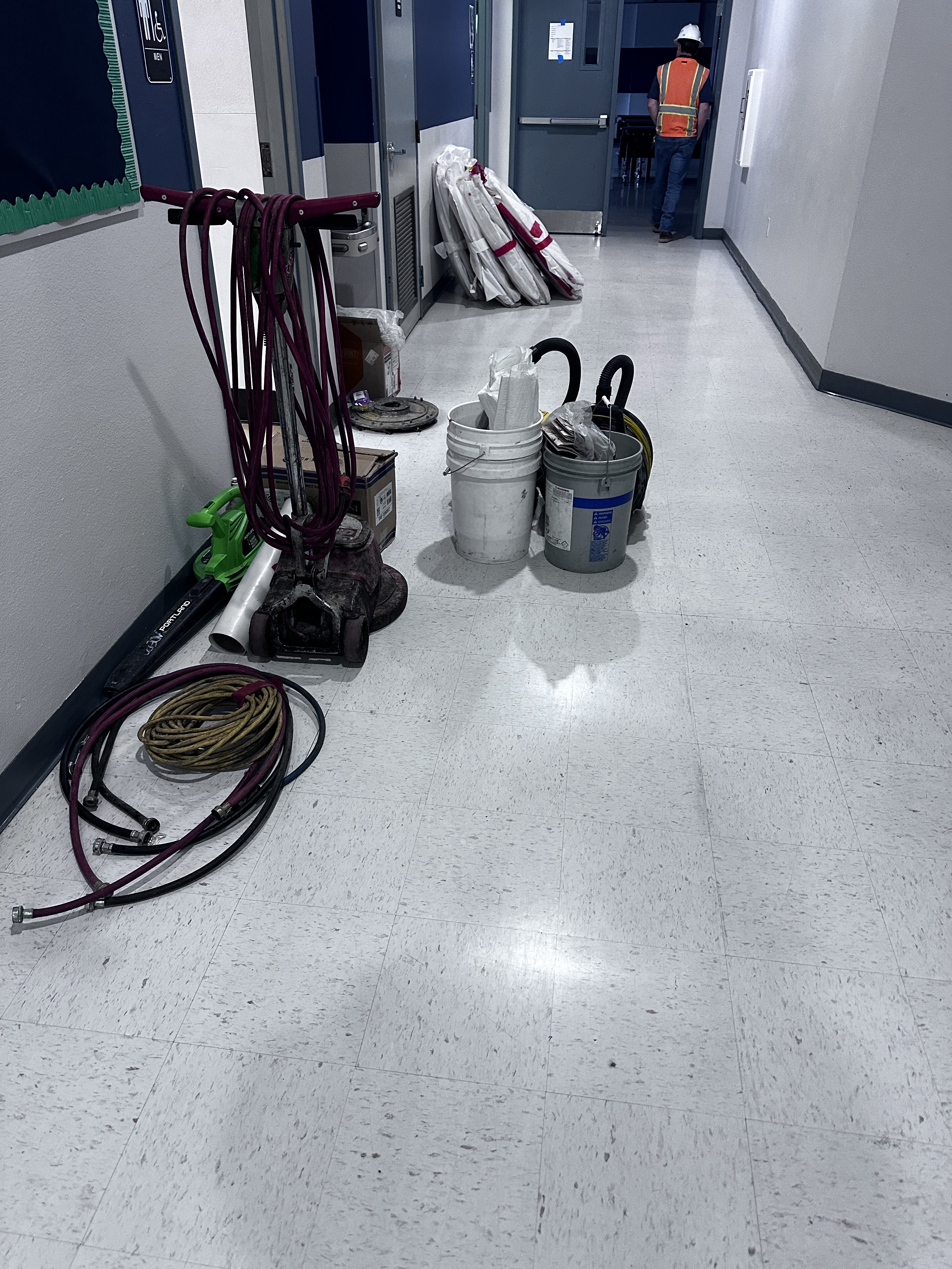 A hallway with construction equipment including hoses, buckets, and a vacuum cleaner, with a person in a safety vest in the background.