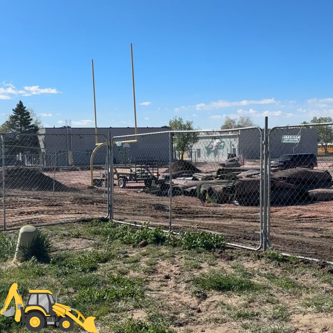 A construction site with a fenced area showing dirt and equipment, including goalposts in the background under a clear blue sky.