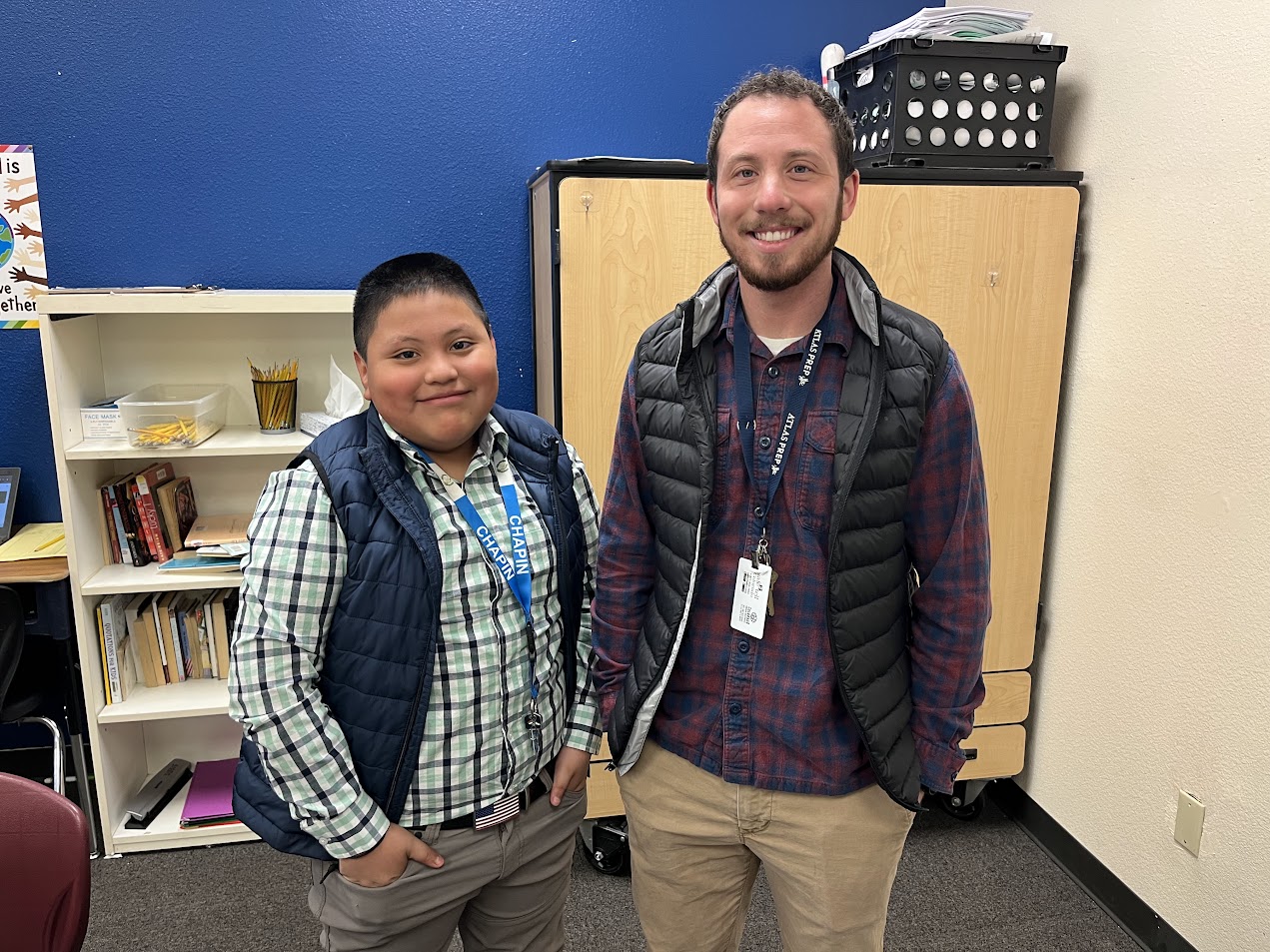 A young boy and a man stand side by side in a classroom, both wearing vests and smiling, with bookshelves and a storage unit in the background.