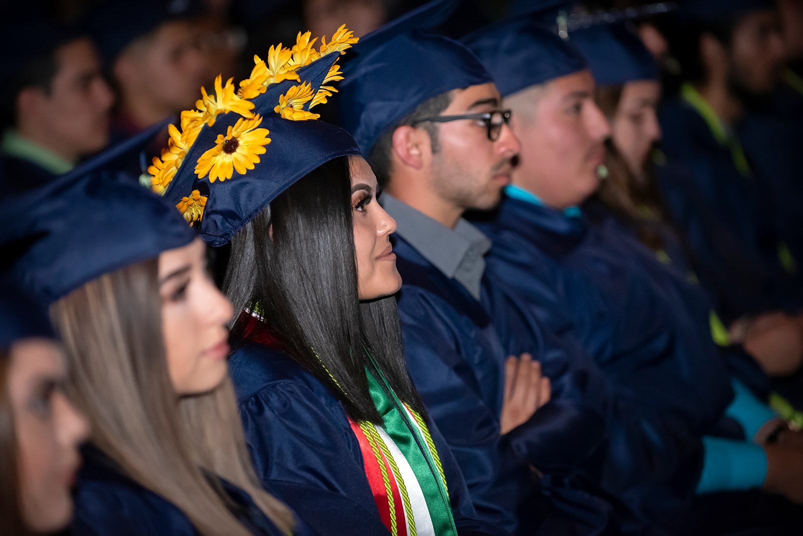A group of graduates seated in a ceremony, with one female graduate wearing a decorated cap adorned with sunflowers, while others are dressed in blue gowns and caps.