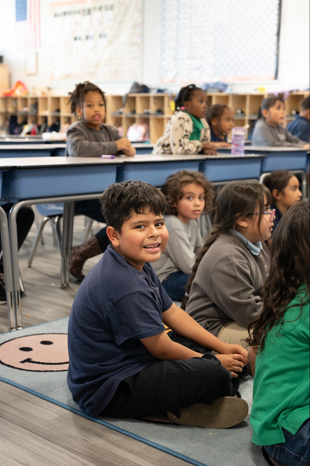 A group of young children sitting on the floor in a classroom, with several raising their hands to participate.