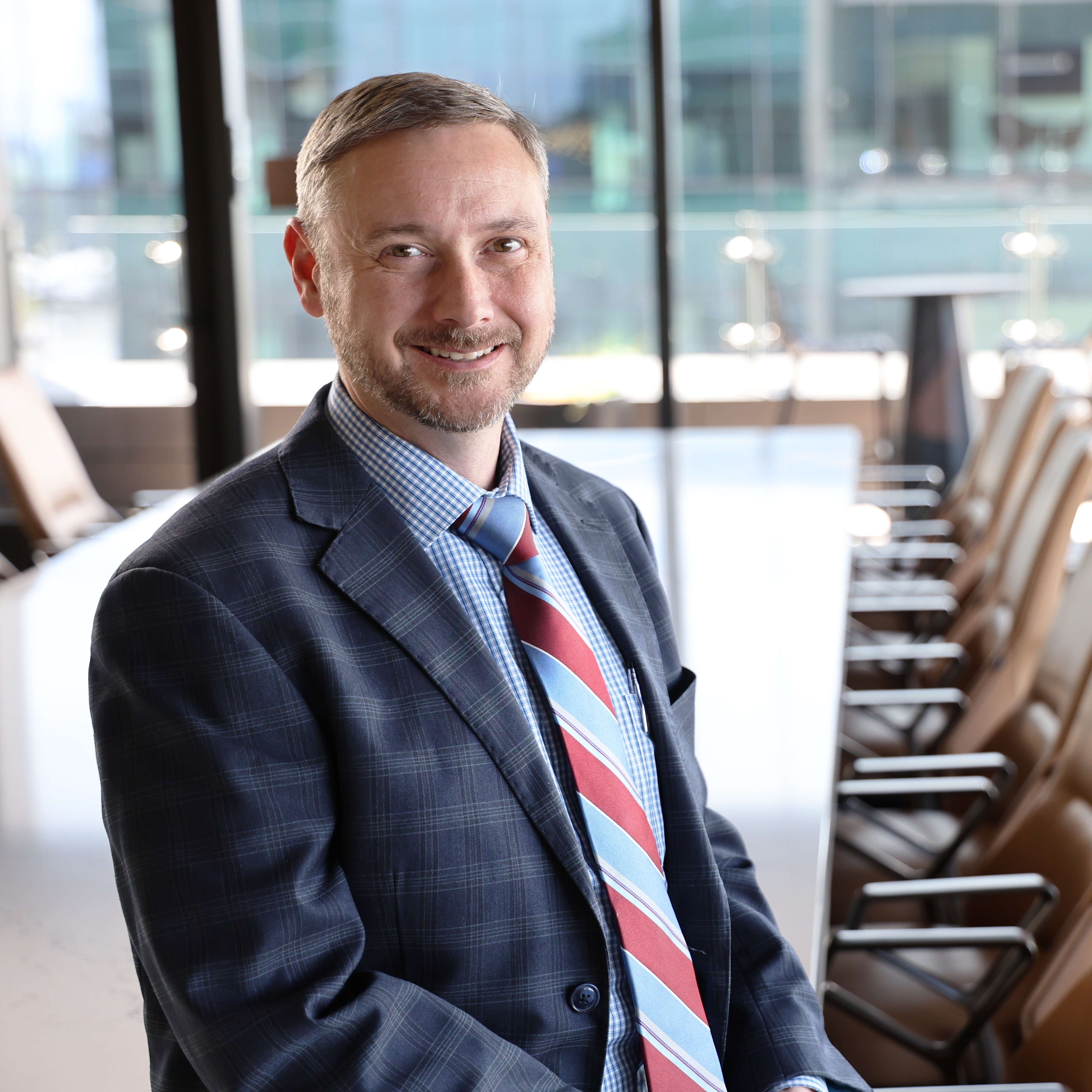 A man with short hair and a beard, wearing a suit and a checkered shirt, smiles at the camera in an office setting with chairs in the background.