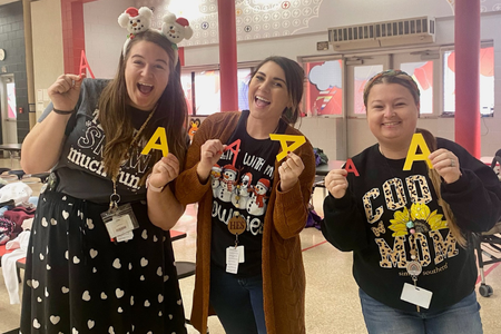 Three smiling educators stand in a school cafeteria holding large yellow letter A’s, celebrating academic success. They are wearing festive clothing, including themed T-shirts and holiday accessories, and appear to be part of a cheerful school spirit event.