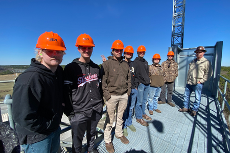 A group of high school students wearing orange hard hats stands on a metal platform during a career and technical education field trip. They are dressed in jackets and jeans, accompanied by an adult supervisor. A large structure and scenic landscape are visible in the background under a clear blue sky.