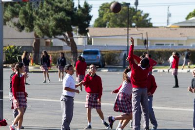 A group of students in red uniforms are playing basketball on a school playground, with some children watching and others participating in the game.
