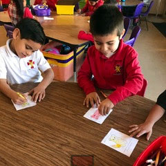 Two young children are seated at a table, focused on coloring small drawings, with various art supplies visible around them.