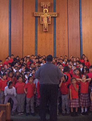 A large group of children in red shirts and plaid skirts stands in a church, facing an adult who is addressing them, with a wooden cross visible in the background.
