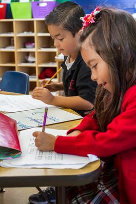 Two children are seated at a table in a classroom, focused on their writing activities, with one girl in a red sweater and a boy in a black sweater, both using pencils on their papers.