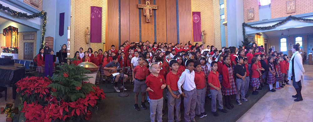 A large group of children in red shirts and gray pants stands together in a church, surrounded by festive decorations, with a cross visible in the background.