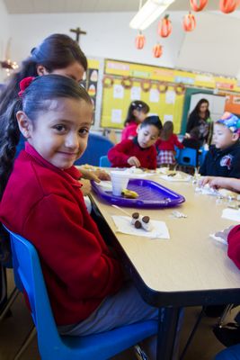A young girl with a red sweater smiles at the camera while sitting at a table with classmates, who are engaged in an activity involving food, in a colorful classroom setting.