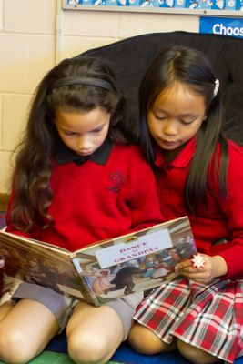 Two young girls in red sweaters sit closely together on a colorful rug, engrossed in reading a book.