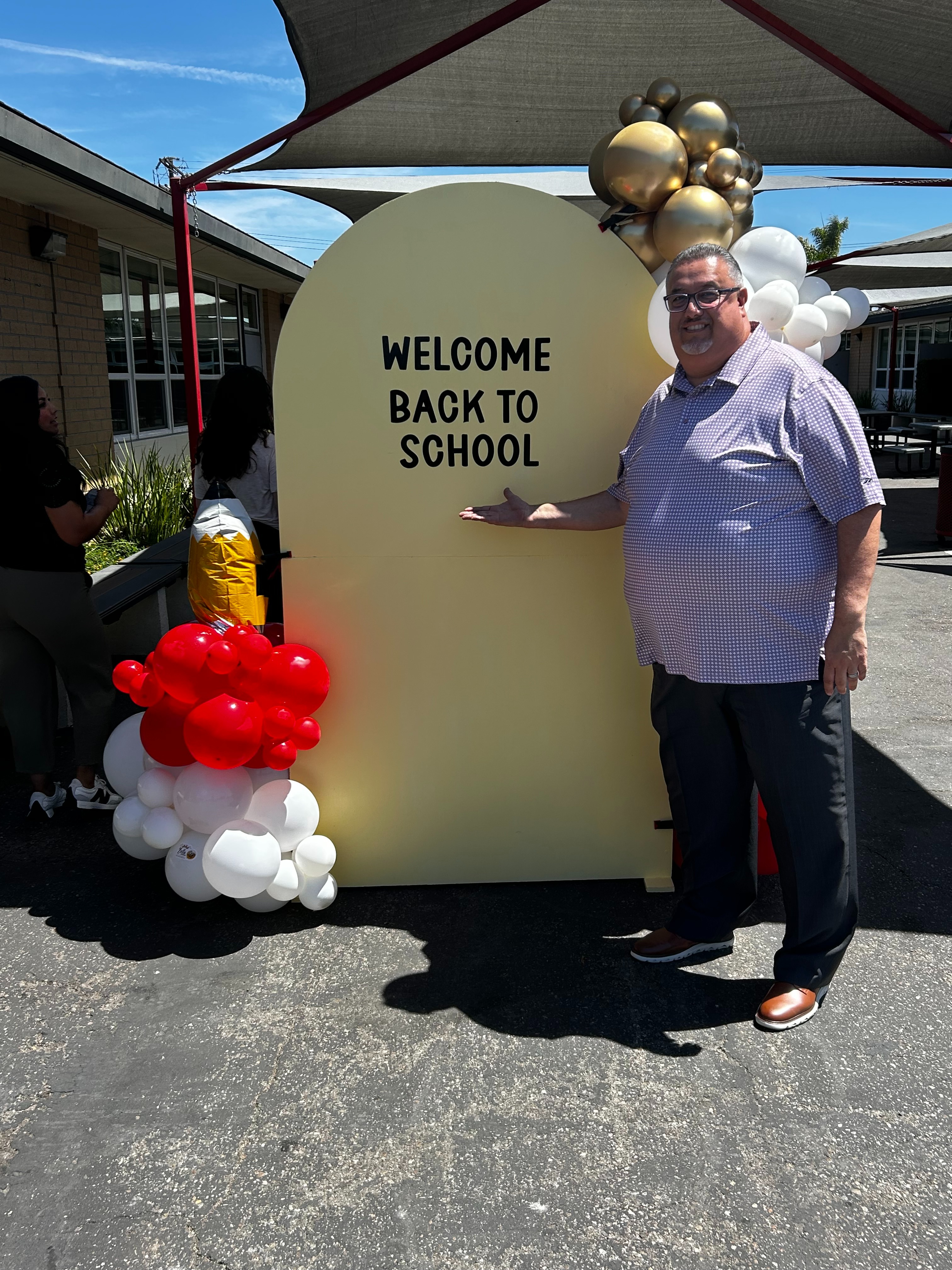 A statue of a standing figure holding a staff, accompanied by a small dog, set against a textured wall and surrounded by lush green plants.