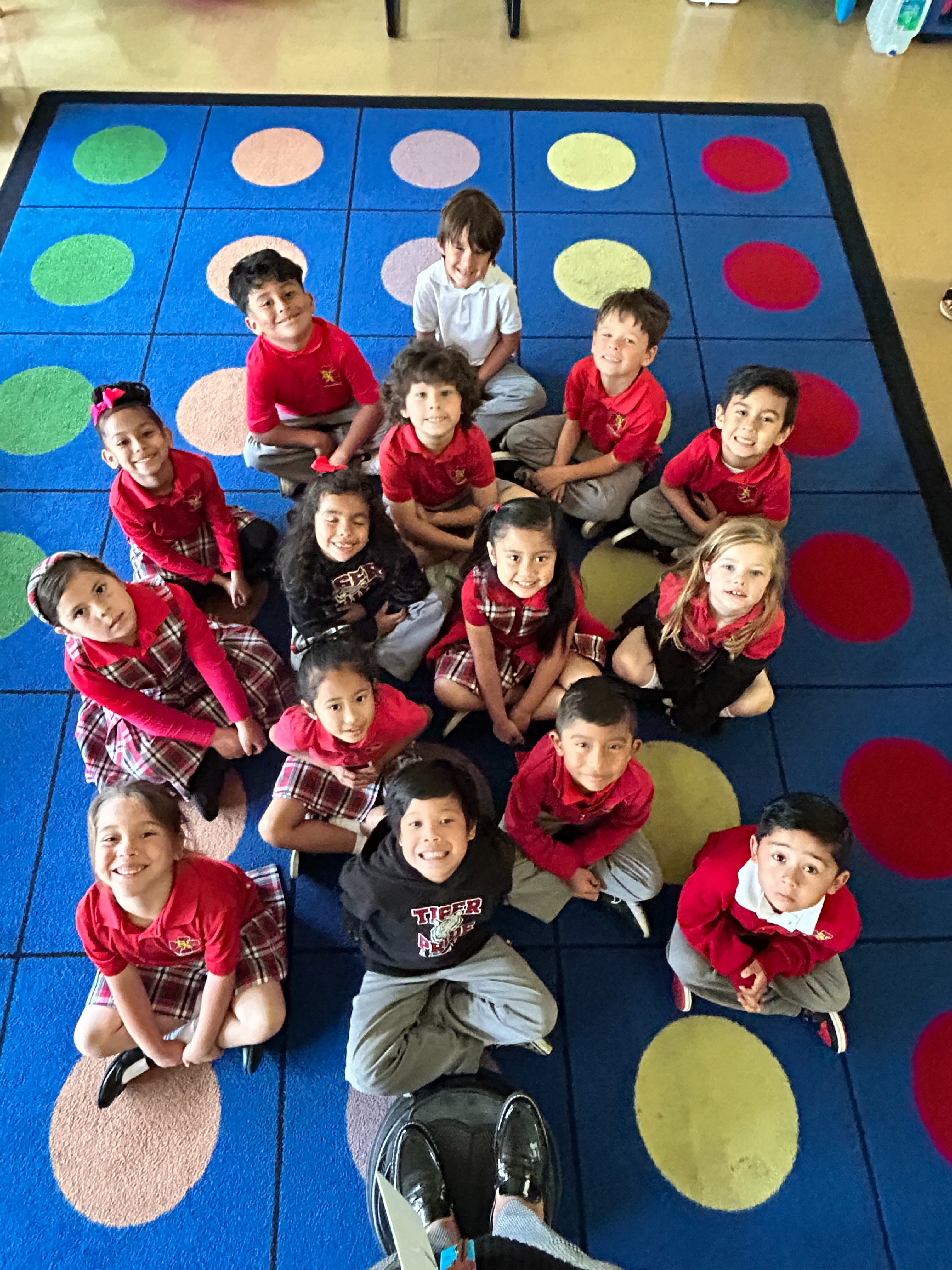 A large group of children in red shirts and plaid skirts stands in a church, facing an adult who is addressing them, with a wooden cross visible in the background.