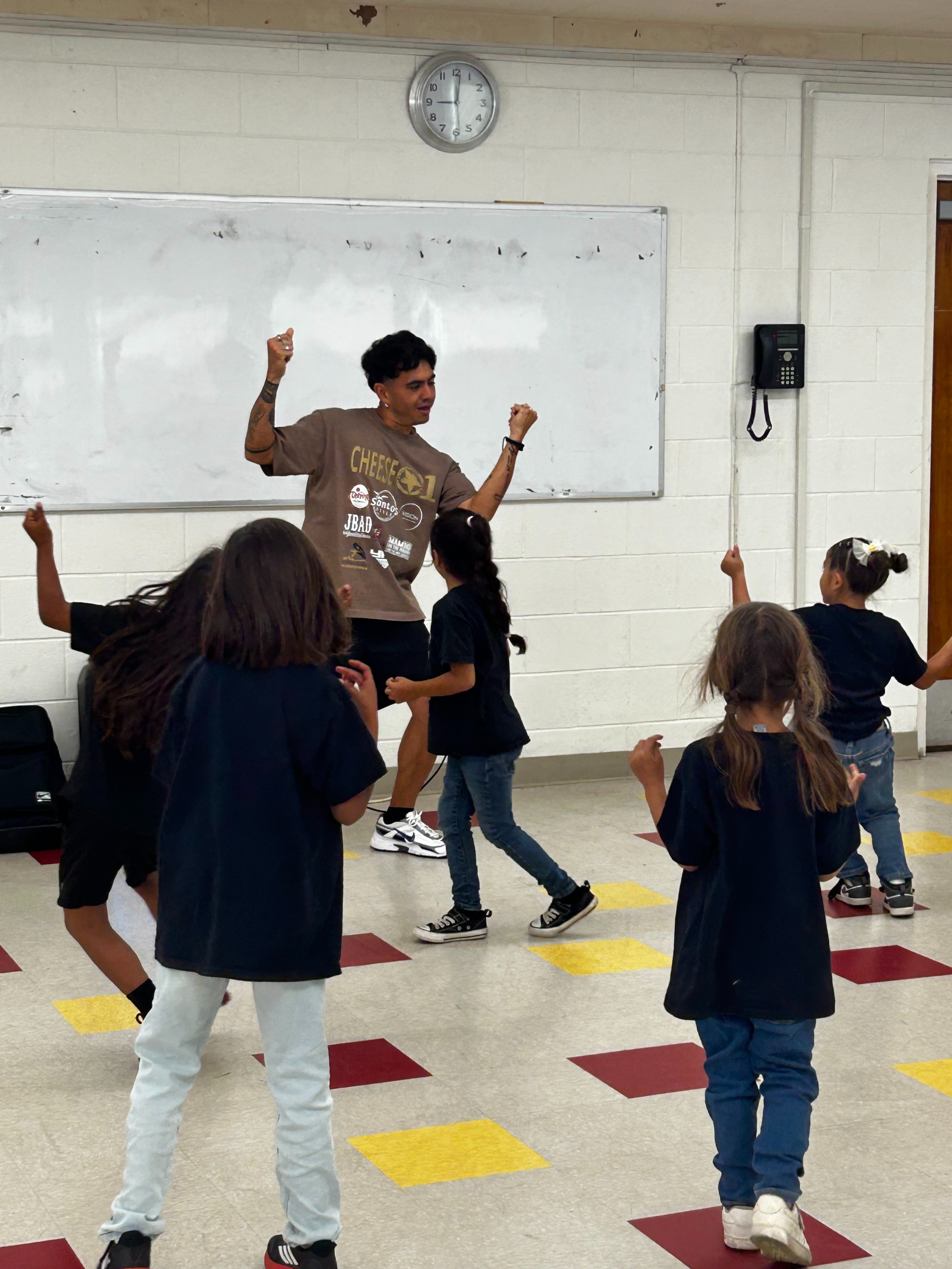 A large group of children in red shirts and plaid skirts stands in a church, facing an adult who is addressing them, with a wooden cross visible in the background.