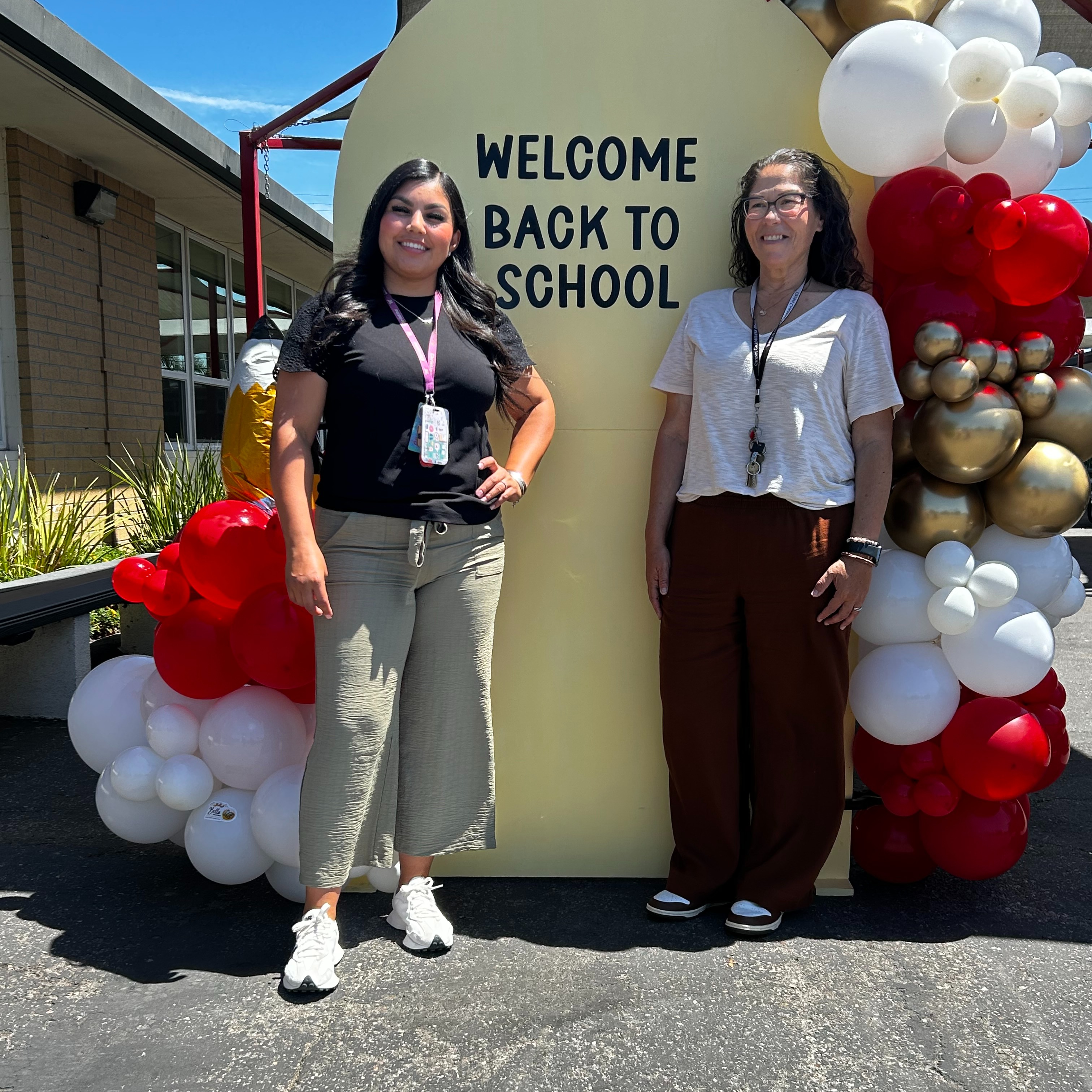 A young girl with a red sweater smiles at the camera while sitting at a table with classmates, who are engaged in an activity involving food, in a colorful classroom setting.