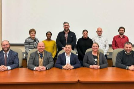 A group of eleven individuals, seated and standing, in a formal setting with a large table, American flags in the background, and a blank screen behind them.