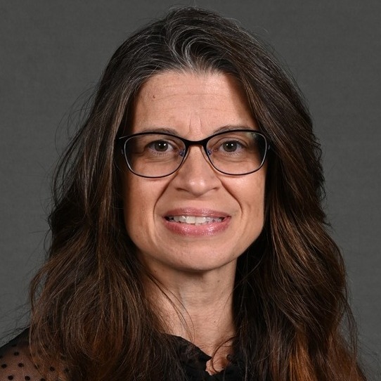 Janet Gesicki wearing a spotted dress shirt with straightened hair and glasses.