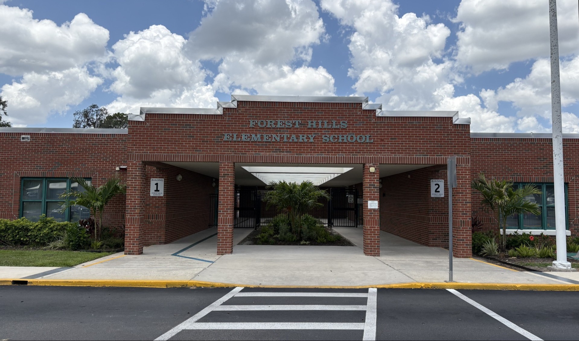 Front entrance of a red-brick school building labeled “Forest Hills Elementary School,” with a covered walkway, crosswalk, and partly cloudy sky overhead.
