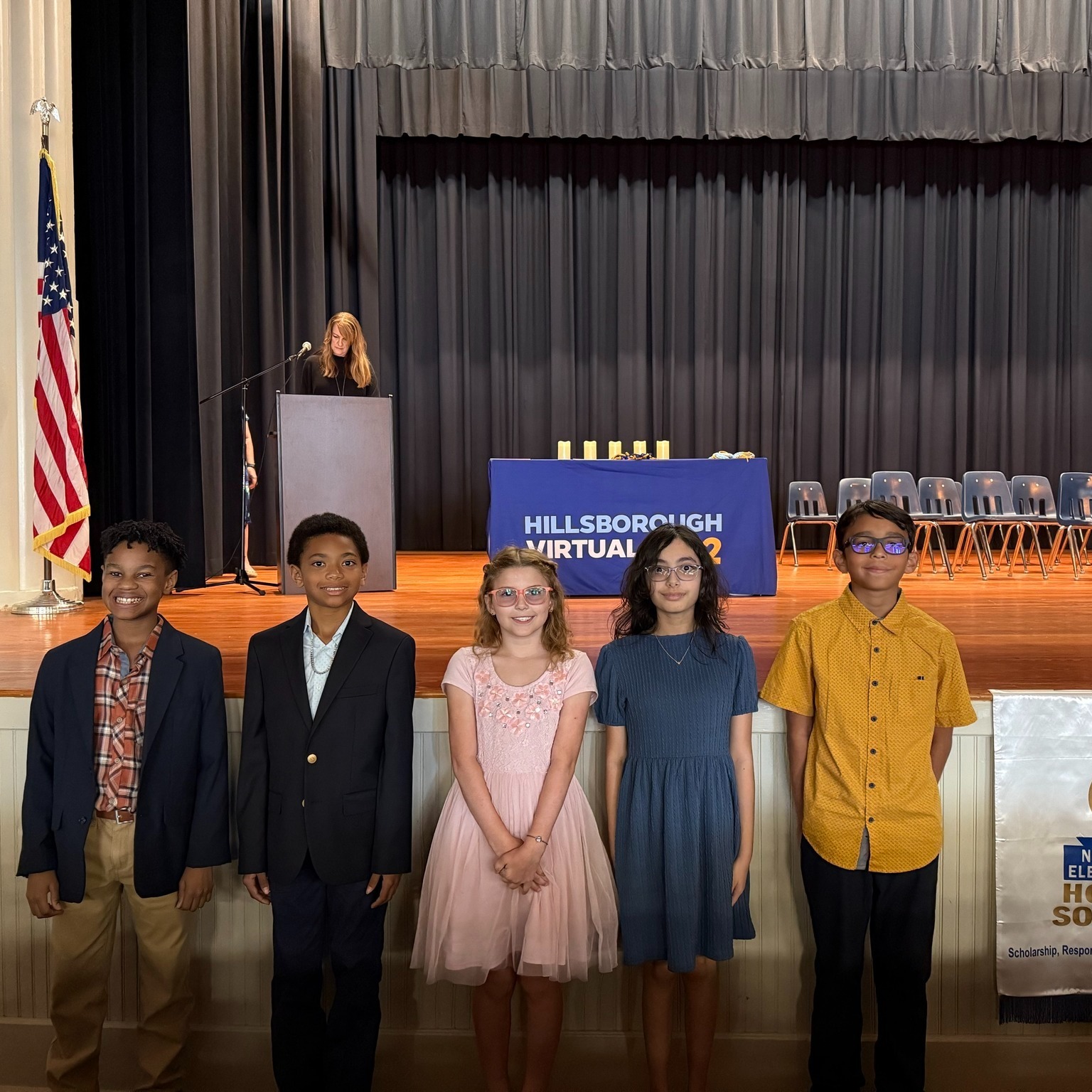 5 elementary students pose in front of the auditorium stage with an administrator on stage at the podium