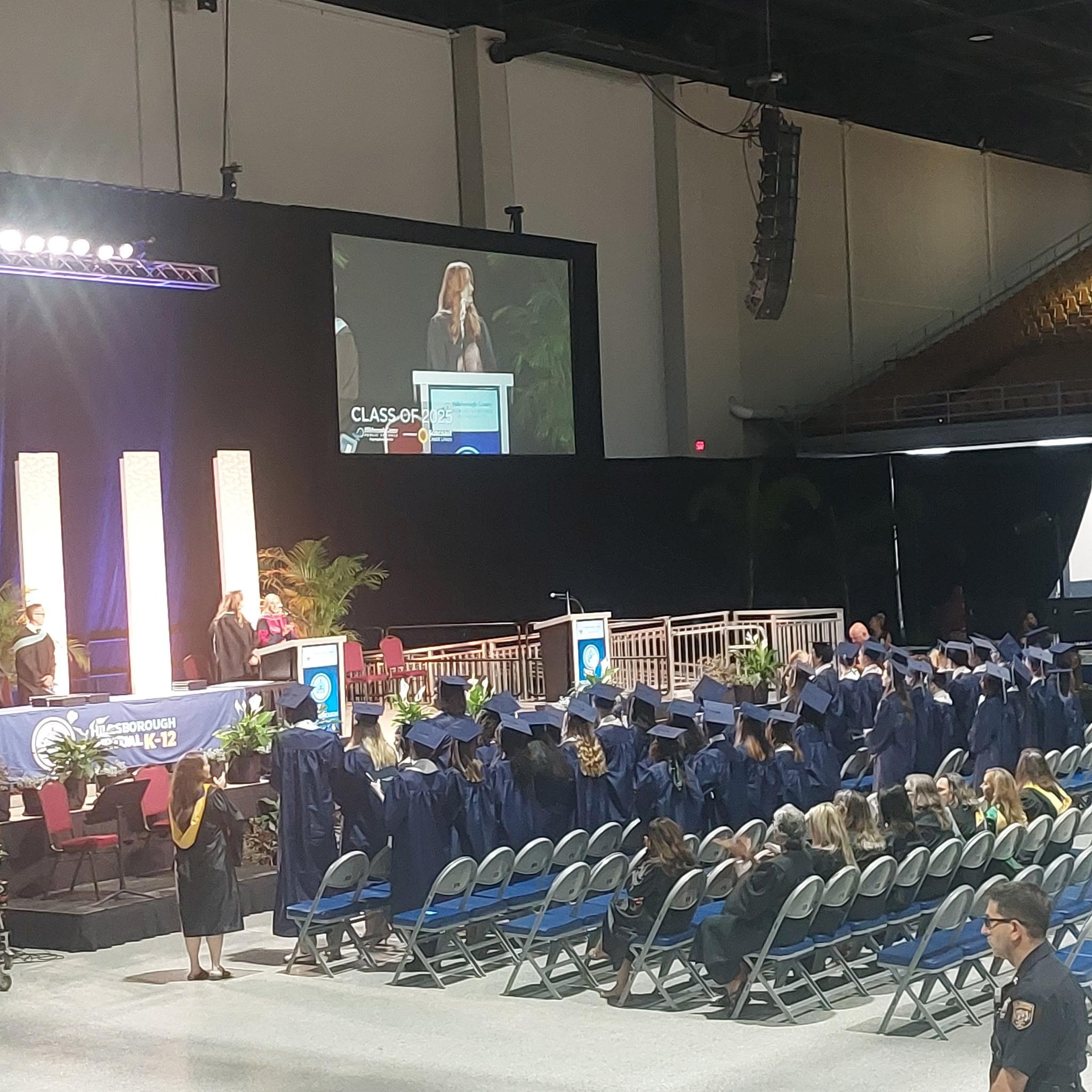 Students in caps and gowns standing up facing graduation podium with 3 administrators standing on stage