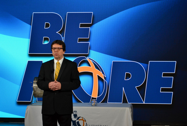 man in black suit speaking in front of blue backdrop with words that say "Be More"