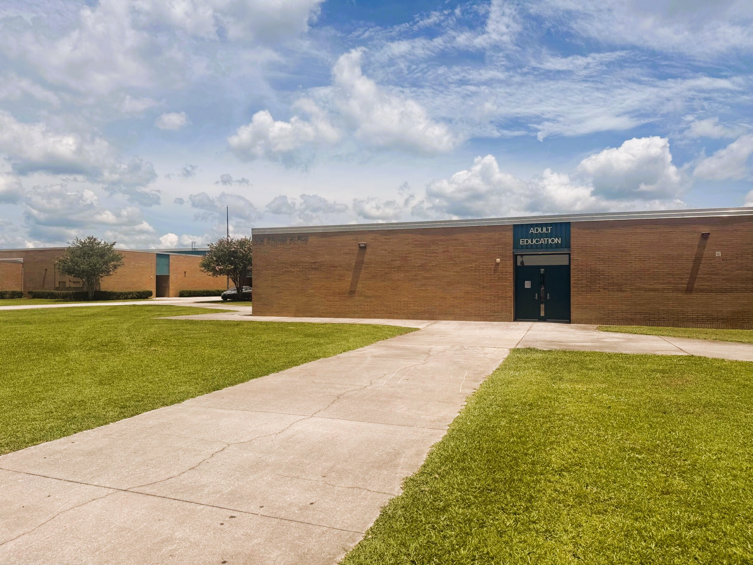 A wide, gently sloped concrete path leads directly to a brown brick building marked "Adult Education" above dark double doors. The entrance is at ground level, providing wheelchair access. The area is open and grassy, with clear visibility and no steps or obstructions. The sky above is partly cloudy with blue patches visible.