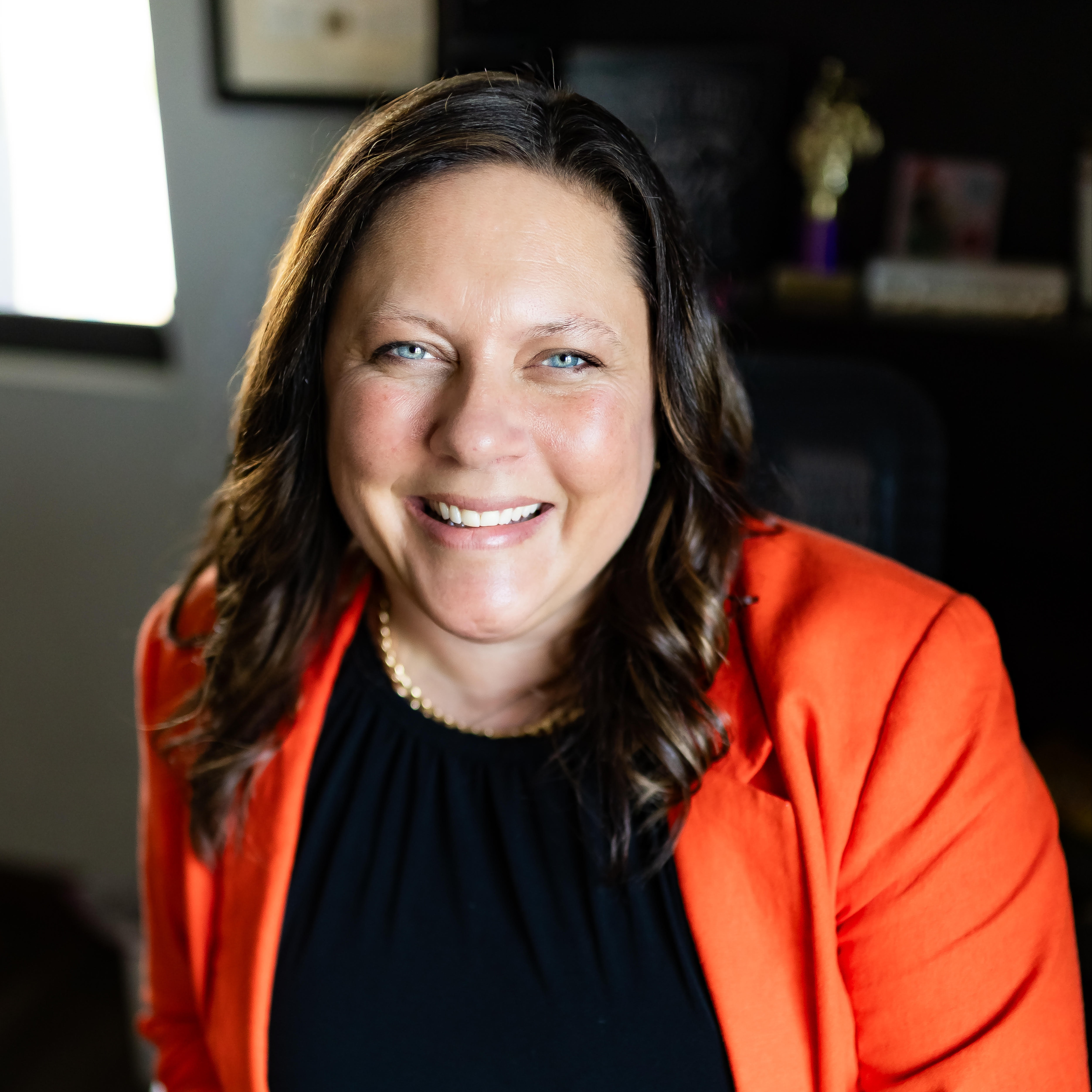 A woman with brown hair sits in a chair, smiling, wearing a black top and orange blazer. Behind her, a window and books are visible.